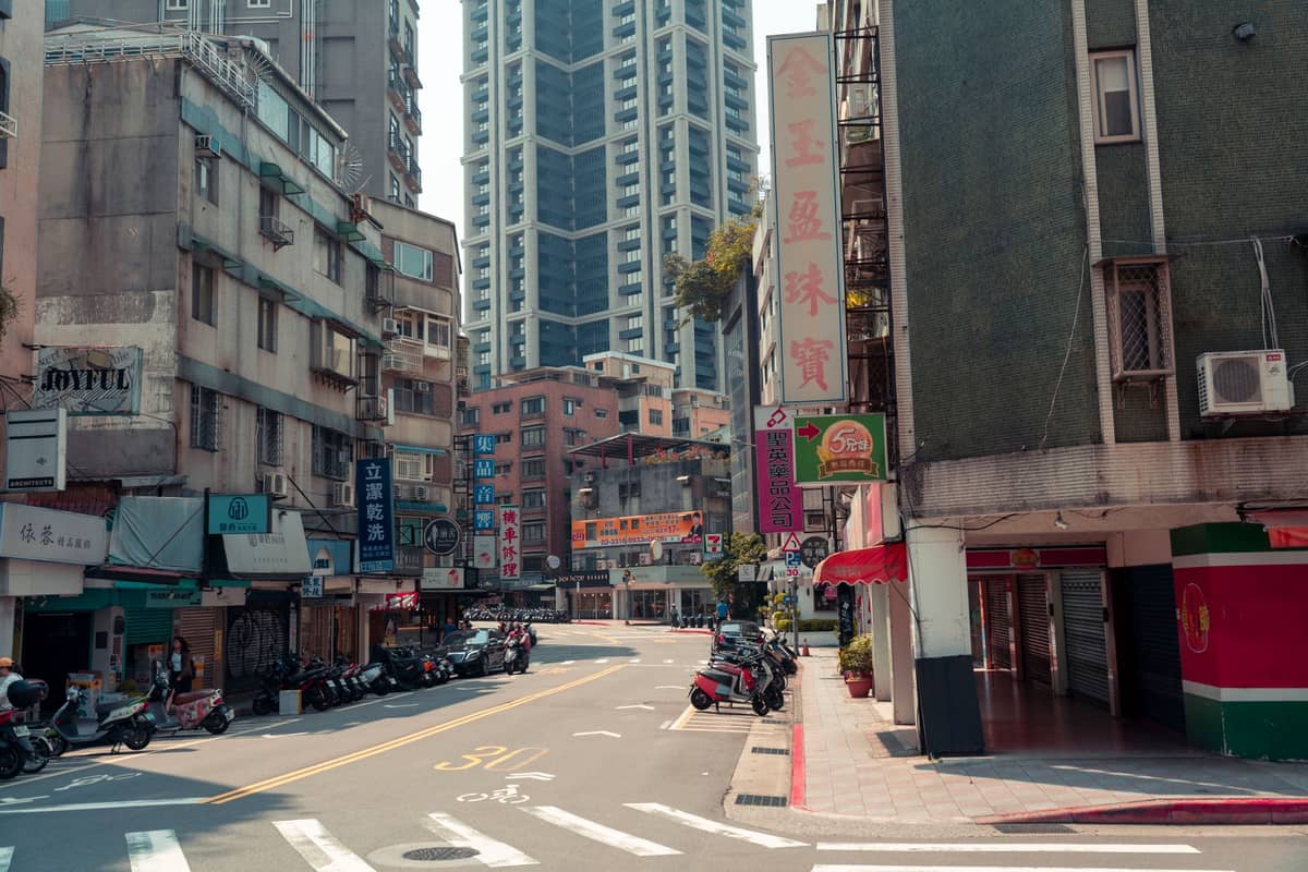 Straßenszene im Zhongshan District in Taipeh mit mittelhohen Wohnhäusern, am Straßenrand geparkten Rollern, chinesischer Beschilderung und Blick auf moderne Hochhäuser im Hintergrund.