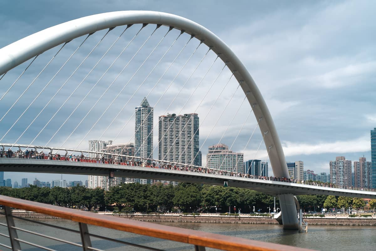 Pedestrian bridge crossing the river in Zhujiang New Town, with modern high-rise buildings and lush green trees under a cloudy sky.