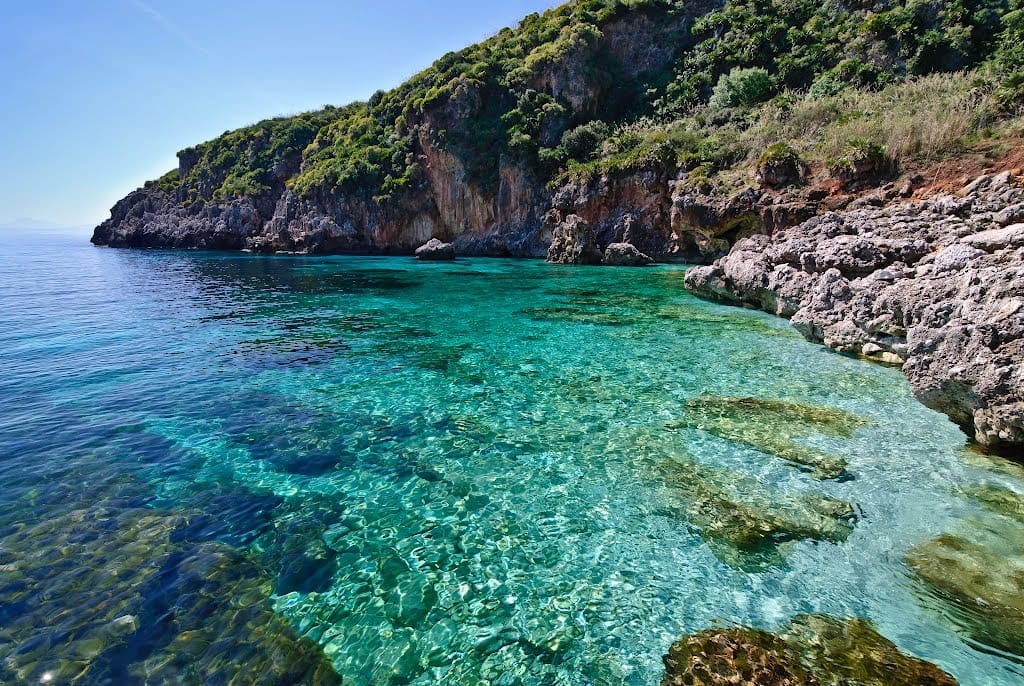 Klares türkisfarbenes Wasser und schroffe Kalksteinklippen entlang der unberührten Küste des Naturschutzgebiets Zingaro auf Sizilien unter einem strahlend blauen Himmel.
