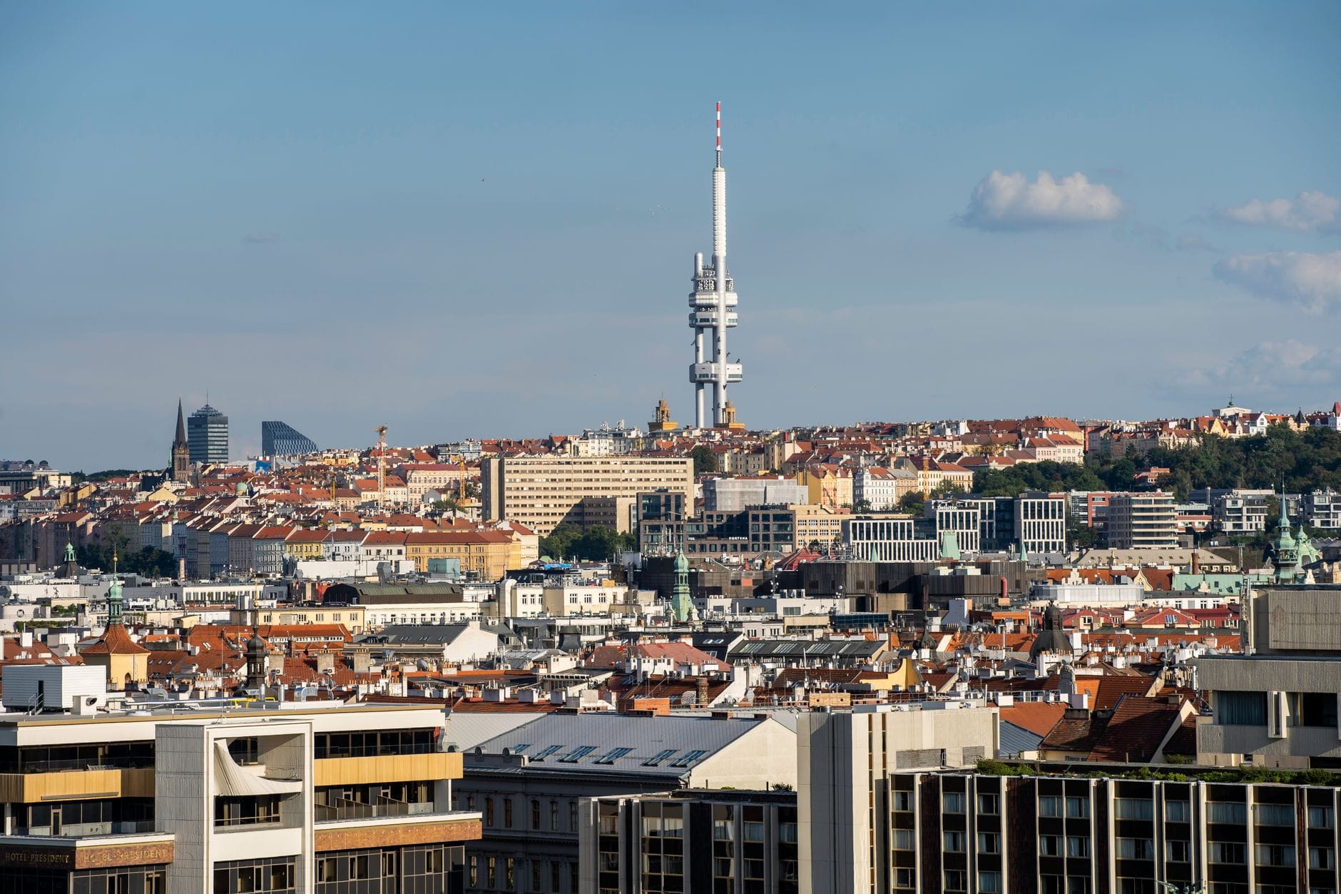 Wieża Telewizyjna Žižkov wznosi się ponad panoramę Pragi w słoneczny dzień, otoczona miejskimi dachami i nowoczesnymi budynkami na pierwszym planie.