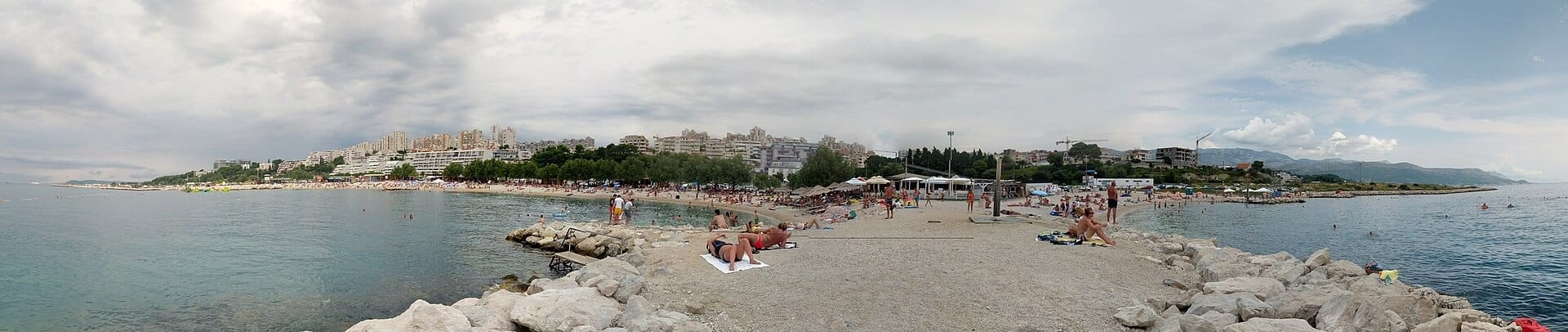 วิวพาโนรามาของหาด Žnjan ในเมือง Split แสดงให้เห็นชายหาดกรวด นักอาบแดด น้ำทะเลสีฟ้าใส และอาคารสมัยใหม่ในพื้นหลัง