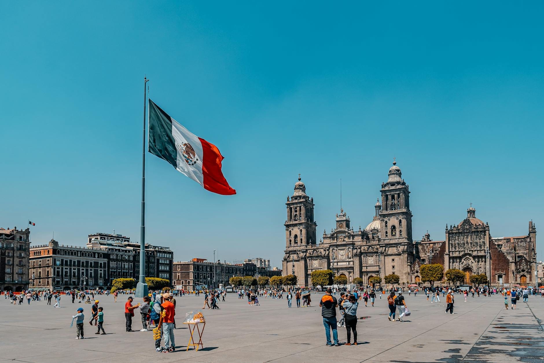 Vista ampia dello Zócalo di Città del Messico con una grande bandiera messicana, persone a passeggio e la Cattedrale Metropolitana sotto un cielo azzurro limpido.
