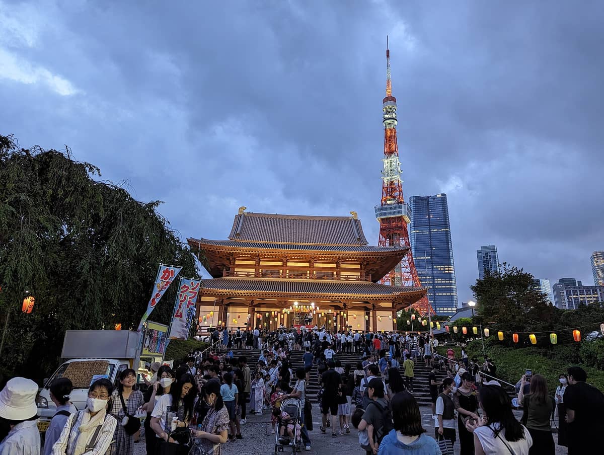 Crowds gathered at Zojo-ji Temple during a festival, with the illuminated Tokyo Tower rising directly behind the temple at dusk in central Tokyo.