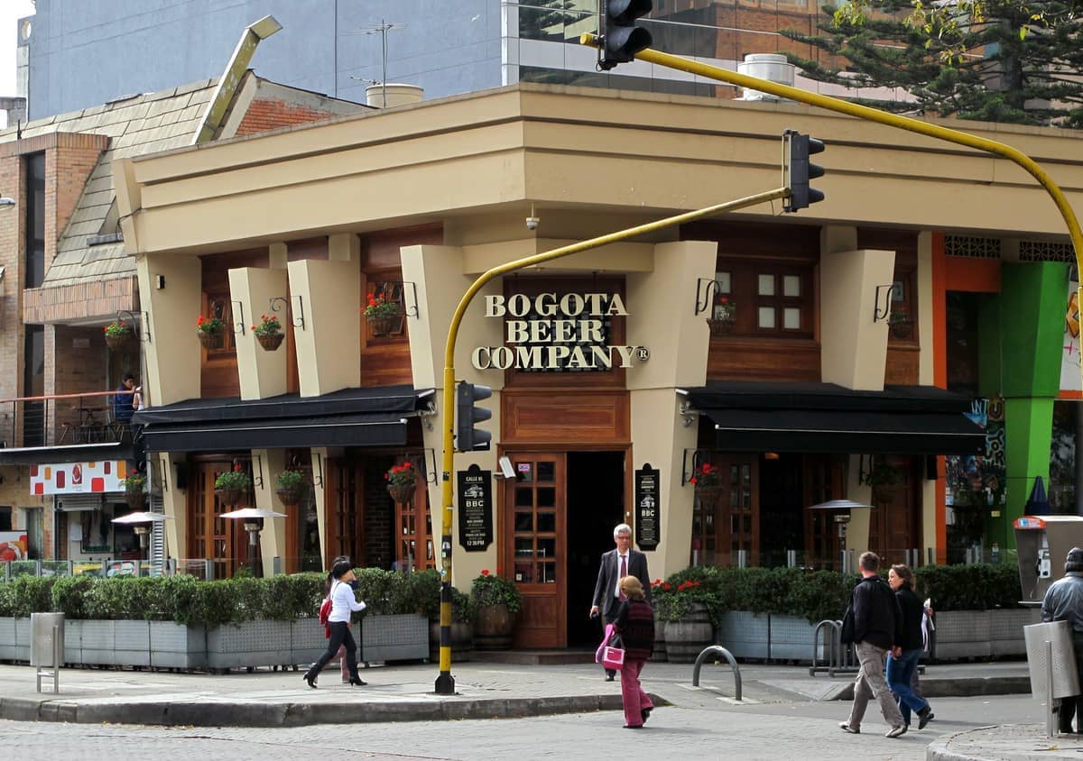 Street view of the Bogota Beer Company building in Zona Rosa, with potted flowers, glass windows, and pedestrians crossing the intersection.