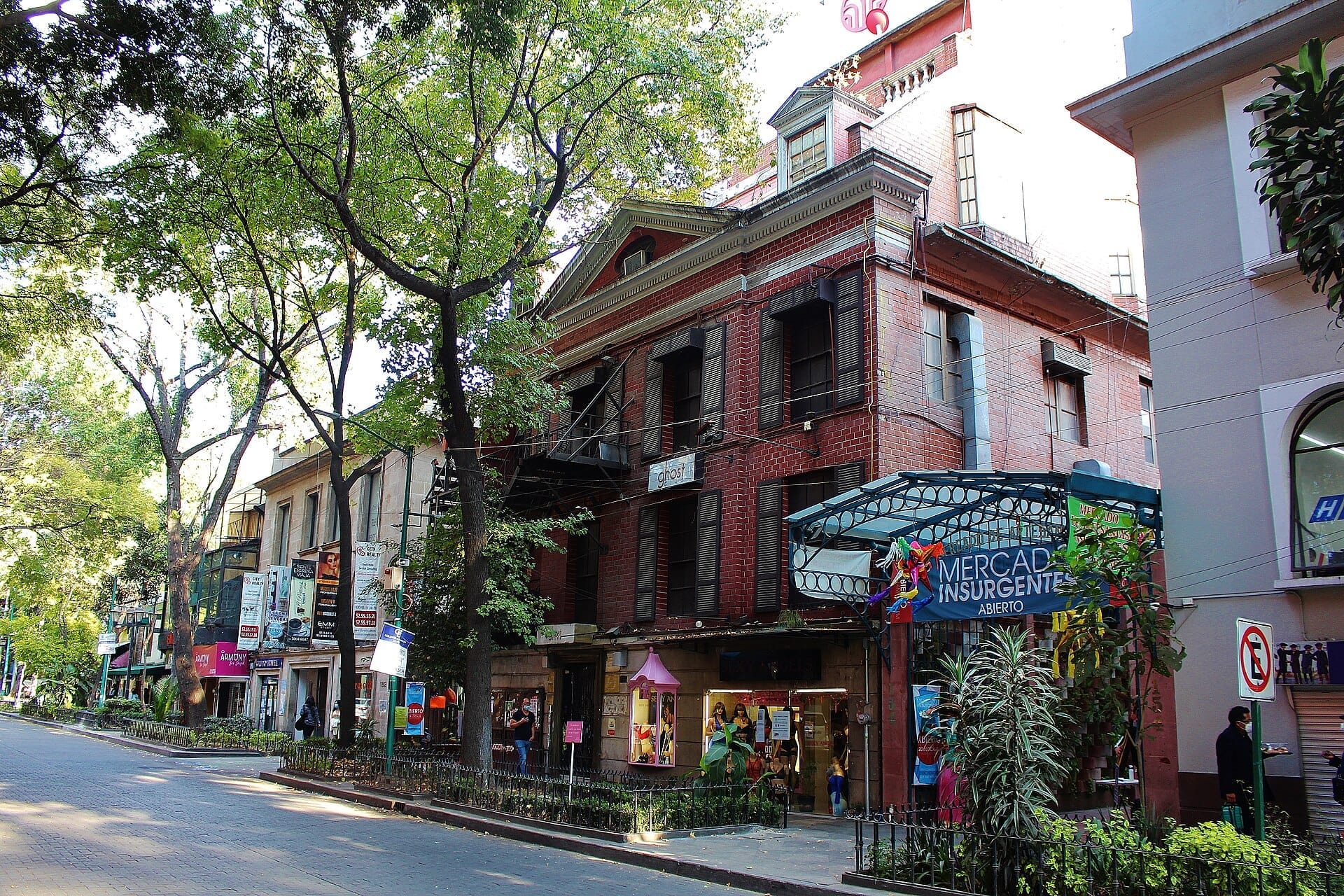 Vista della strada della Zona Rosa a Mexico City con storici edifici in mattoni, alberi frondosi e l'ingresso al Mercado Insurgentes in una giornata luminosa.