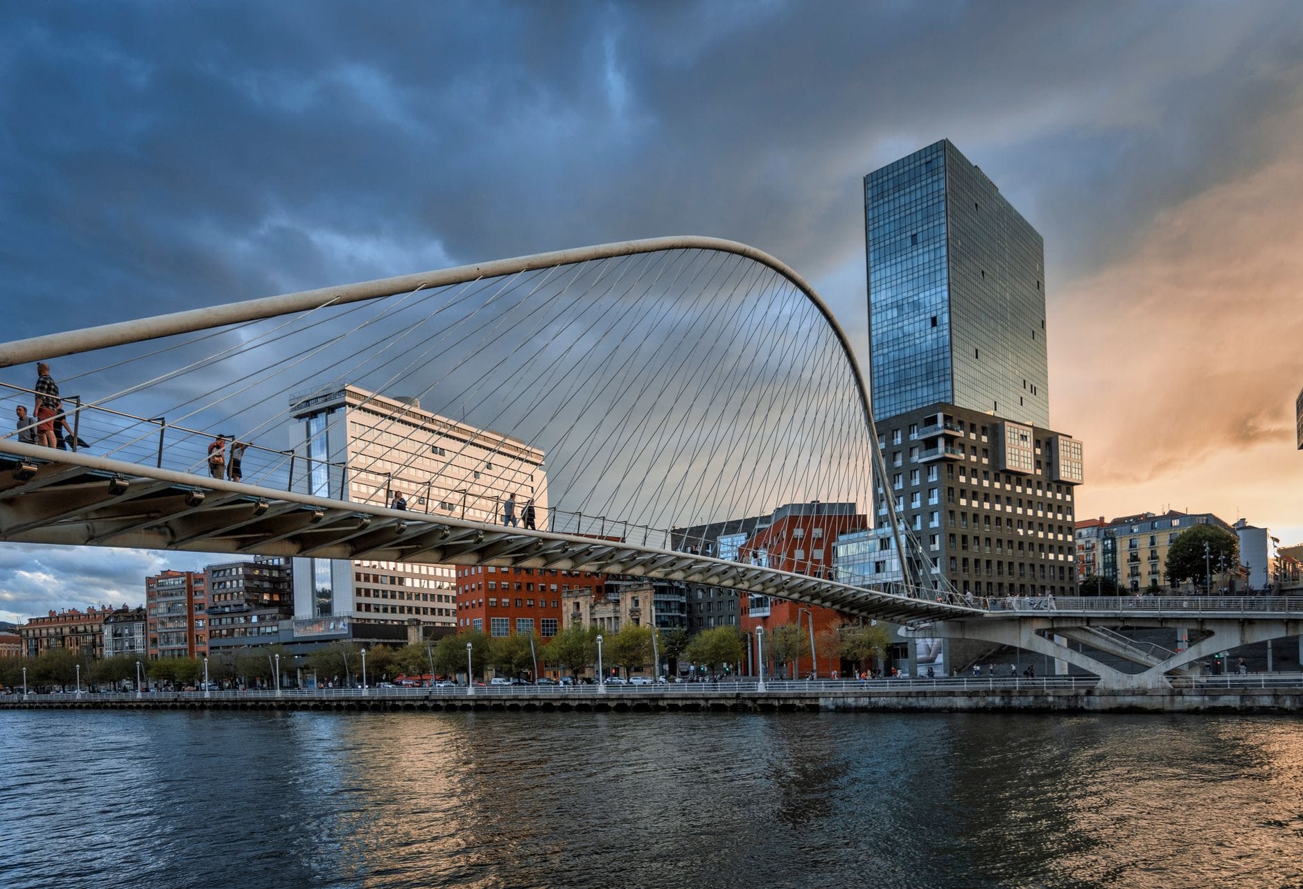 The Zubizuri Bridge in Bilbao dramatically spanning the Nervión River at sunset, with people crossing and modern city buildings in the background.