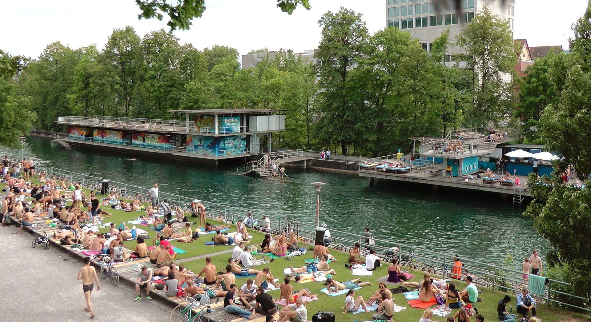 ผู้คนจำนวนมากนอนอาบแดดและพักผ่อนบนริมหญ้าริม Flussbad Oberer Letten สระว่ายน้ำแม่น้ำที่ซูริค มีแพว่ายน้ำ ต้นไม้ และแม่น้ำให้เห็นในภาพ