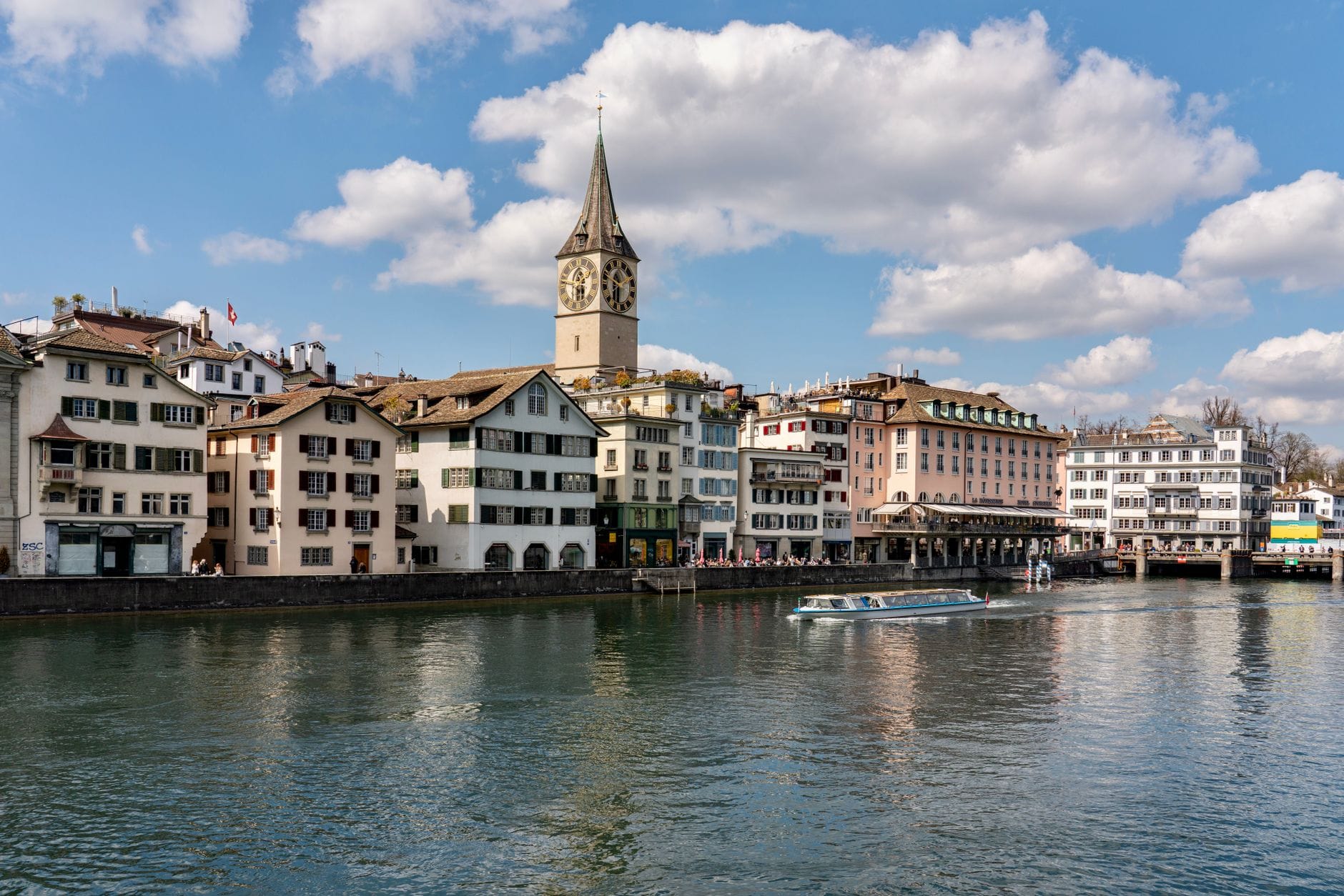 วิวกว้างของโบสถ์ St. Peter พร้อมหอนาฬิกาขนาดใหญ่ริมแม่น้ำในเมือง Zurich ล้อมรอบด้วยอาคารประวัติศาสตร์และสะท้อนอยู่บนผิวน้ำที่เงียบสงบ