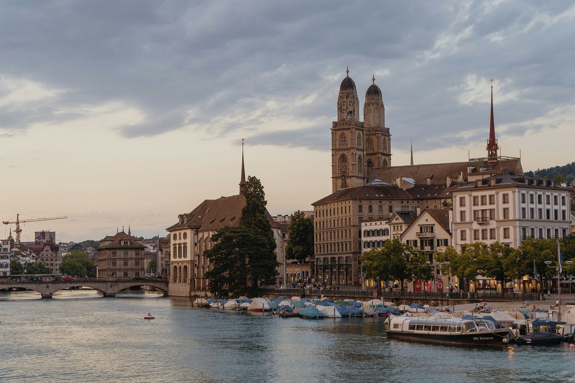 โบสถ์ Grossmünster ในเมืองซูริกพร้อมหอคอยคู่ มองข้ามแม่น้ำ Limmat ในยามพระอาทิตย์ตก ล้อมรอบด้วยอาคารประวัติศาสตร์ของเมือง