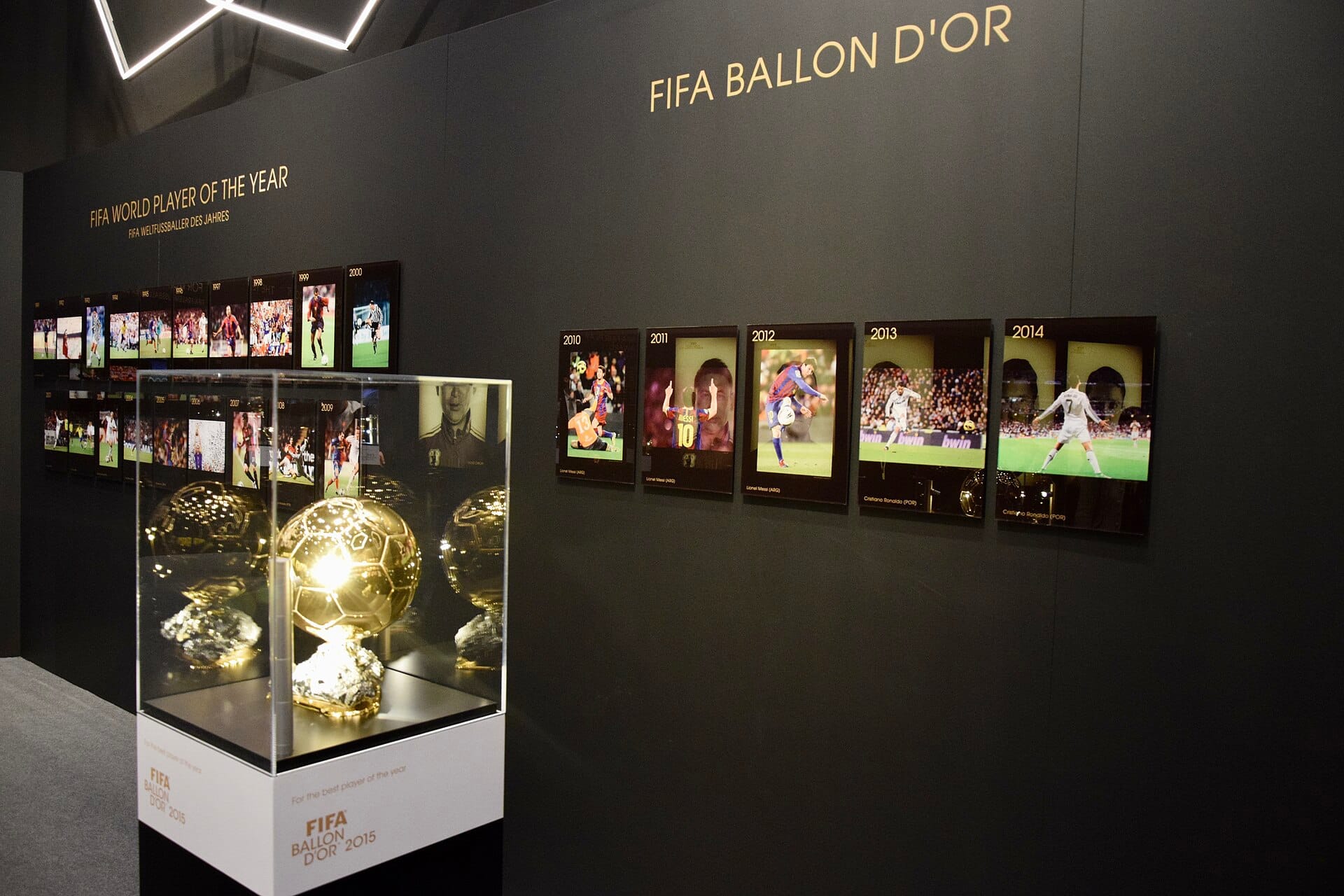 นิทรรศการที่พิพิธภัณฑ์ฟีฟ่า ซูริค โชว์ถ้วยรางวัล FIFA Ballon d'Or อยู่ในตู้กระจก พร้อมภาพอดีตเจ้าของรางวัลบนผนังสีเข้ม