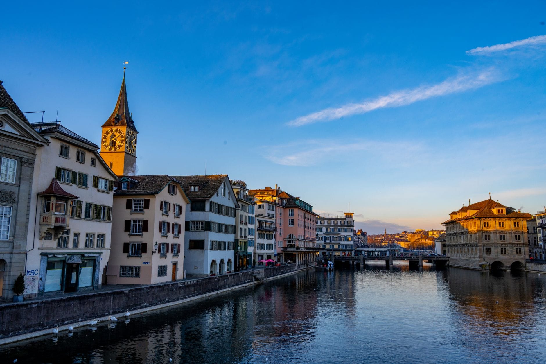 เมืองเก่าซูริคและแม่น้ำ Limmat ยามพระอาทิตย์ตก ตึกประวัติศาสตร์และหอนาฬิกาโบสถ์ St. Peter ริมน้ำ ใต้ท้องฟ้าสีคราม