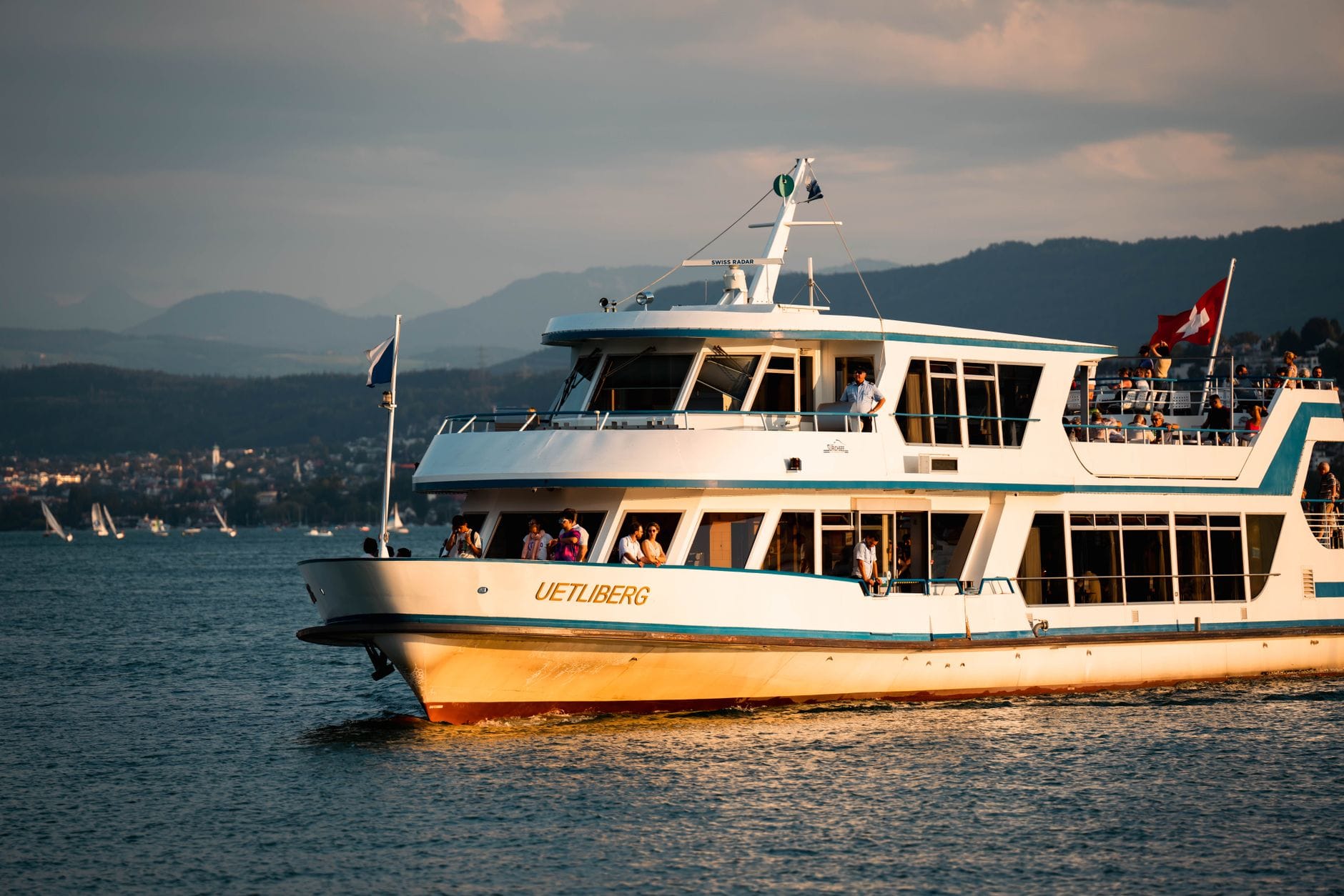 เรือโดยสารชื่อ Uetliberg แล่นอยู่บนทะเลสาบซือริชในช่วงพระอาทิตย์ตก โดยมีผู้โดยสารเพลิดเพลินกับวิวเมืองและเนินเขาเป็นฉากหลัง