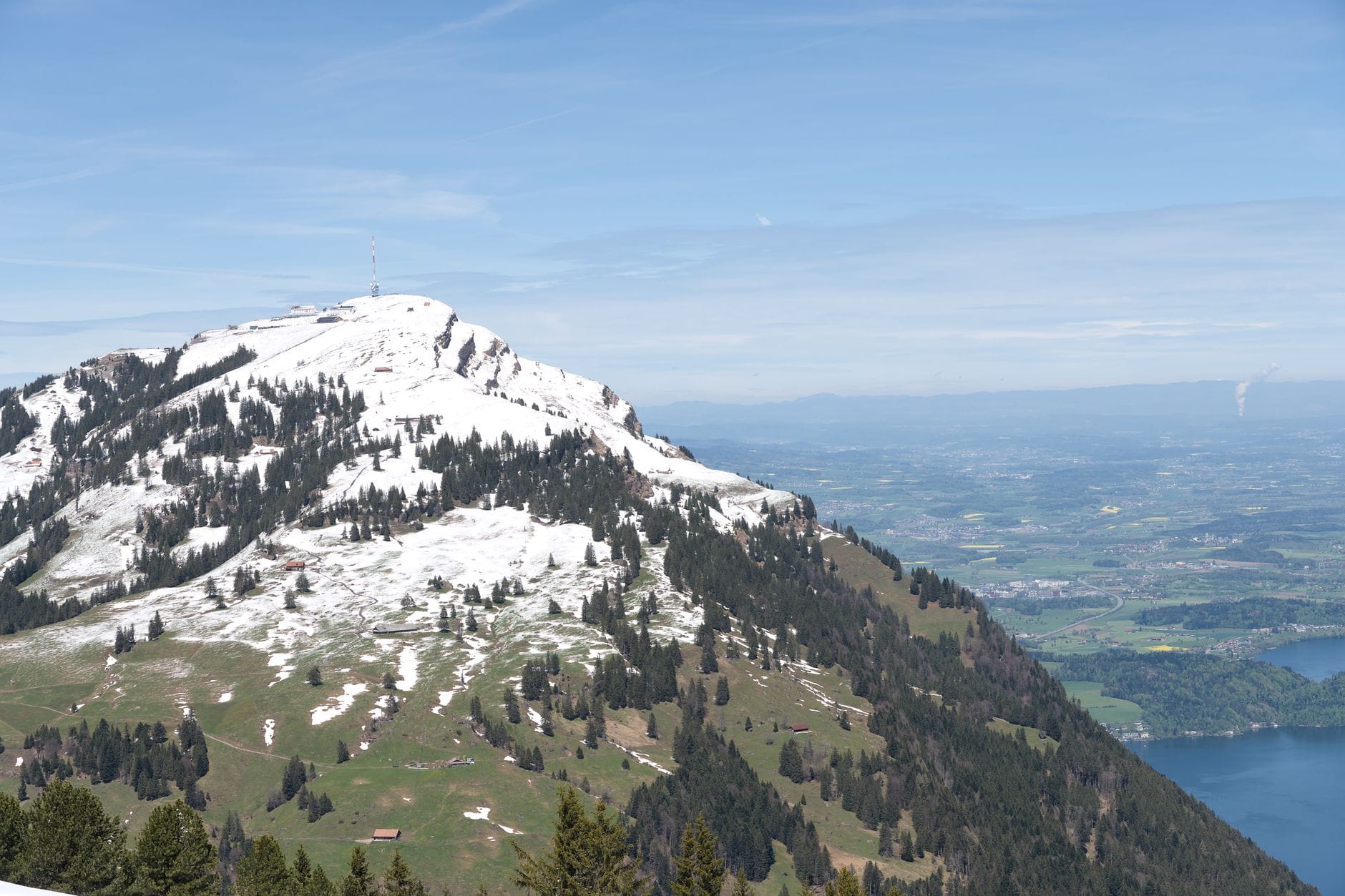 วิวยอดเขา Mount Rigi ที่ปกคลุมด้วยหิมะ ล้อมรอบด้วยเนินเขาสีเขียว ป่าไม้ และทะเลสาบในระยะไกล ใต้ท้องฟ้าสีฟ้าใส