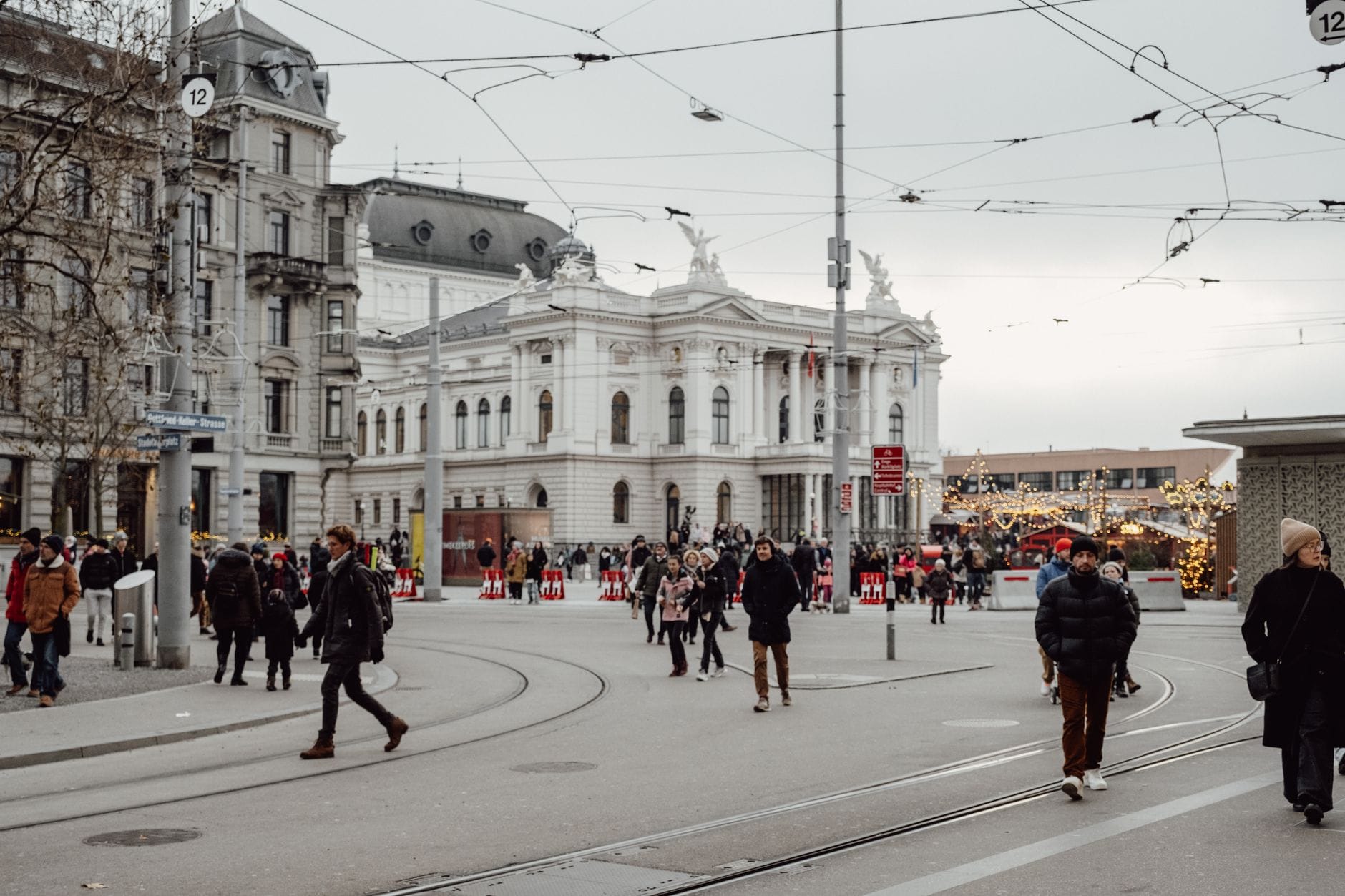 ผู้คนกำลังเดินและรถรางวิ่งอยู่ใกล้โรงอุปรากร Zurich โดยมีแผงตลาดคริสต์มาสอยู่เบื้องหลัง