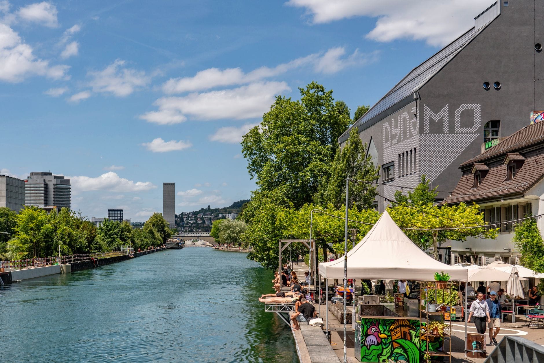 ฉากฤดูร้อนกลางแจ้งริมแม่น้ำ Limmat ในเมือง Zurich พร้อมผู้คนที่เพลิดเพลินกับคาเฟ่และทางเดินริมน้ำ ต้นไม้ร่มรื่น และท้องฟ้าสีฟ้า