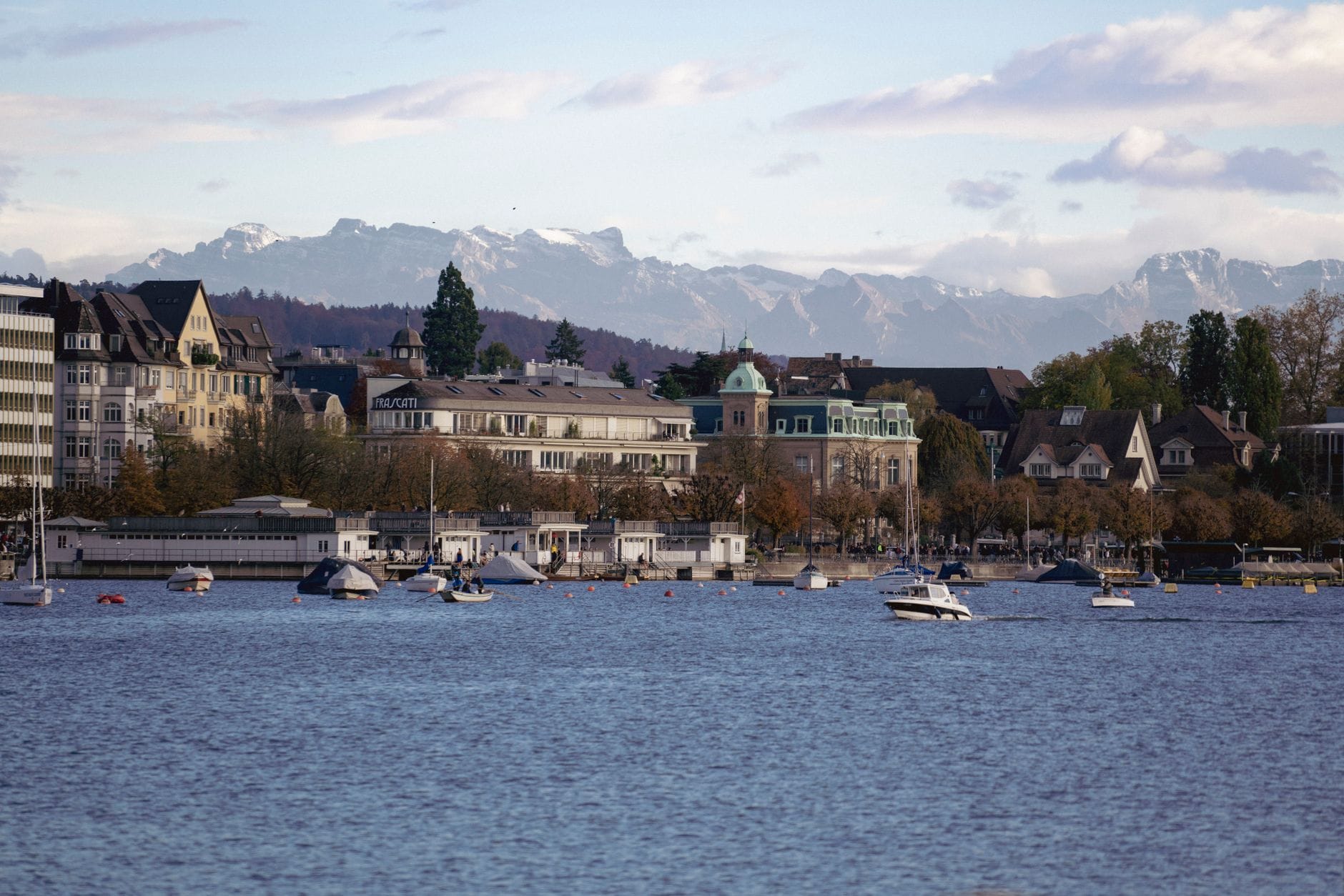 วิวข้ามทะเลสาบซูริค (Lake Zurich) ไปยังชานเมืองริมทะเลสาบที่มีเรือ อาคารที่พักอาศัย และภูเขาในระยะไกลให้เห็นใต้ท้องฟ้าสีฟ้า