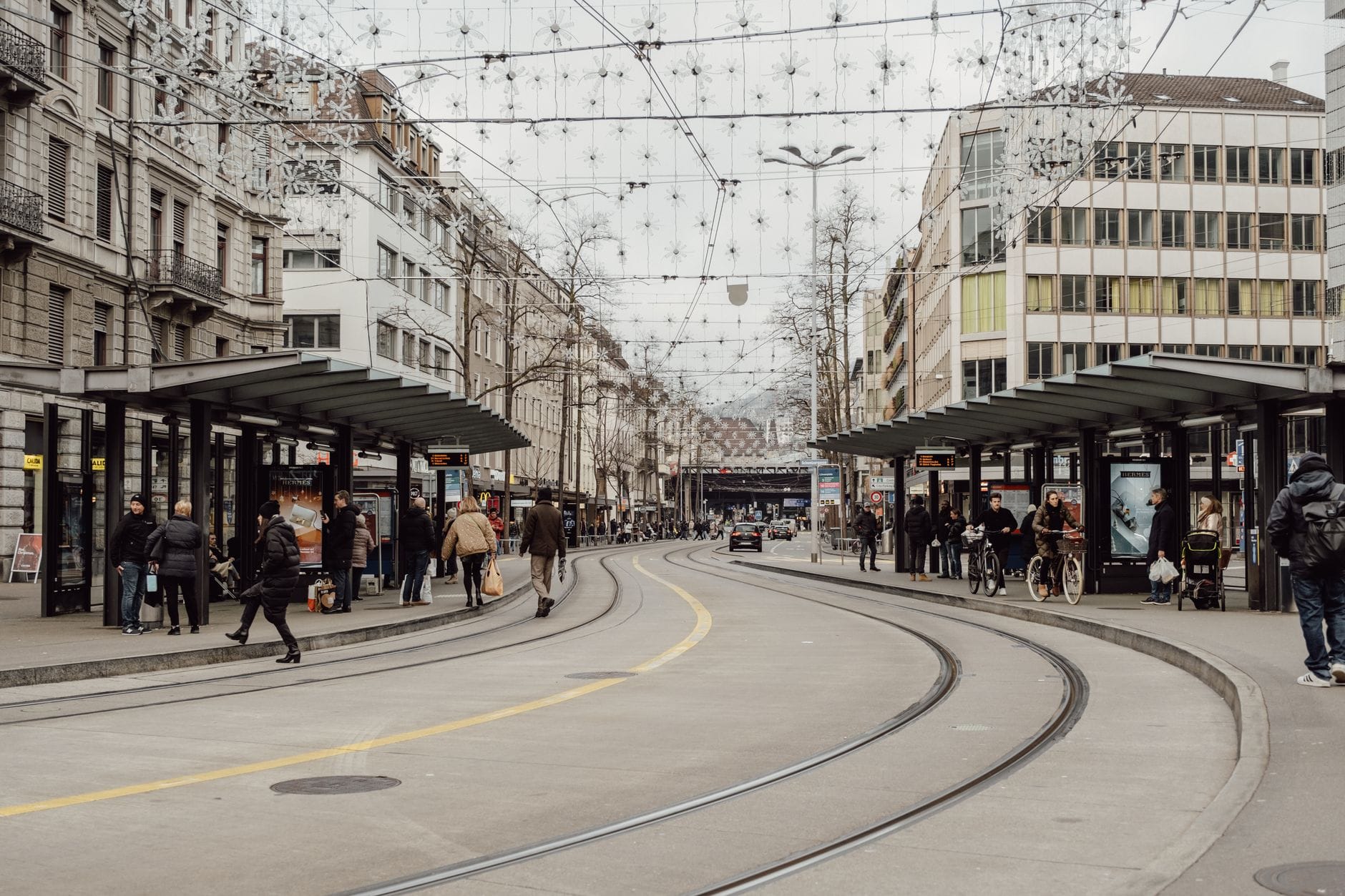 สถานีรถรางที่คึกคักใน Zurich ยามพลบค่ำ มีผู้คนรอรถ ไฟประดับเหนือถนน และอาคารคลาสสิกของเมืองล้อมรอบ