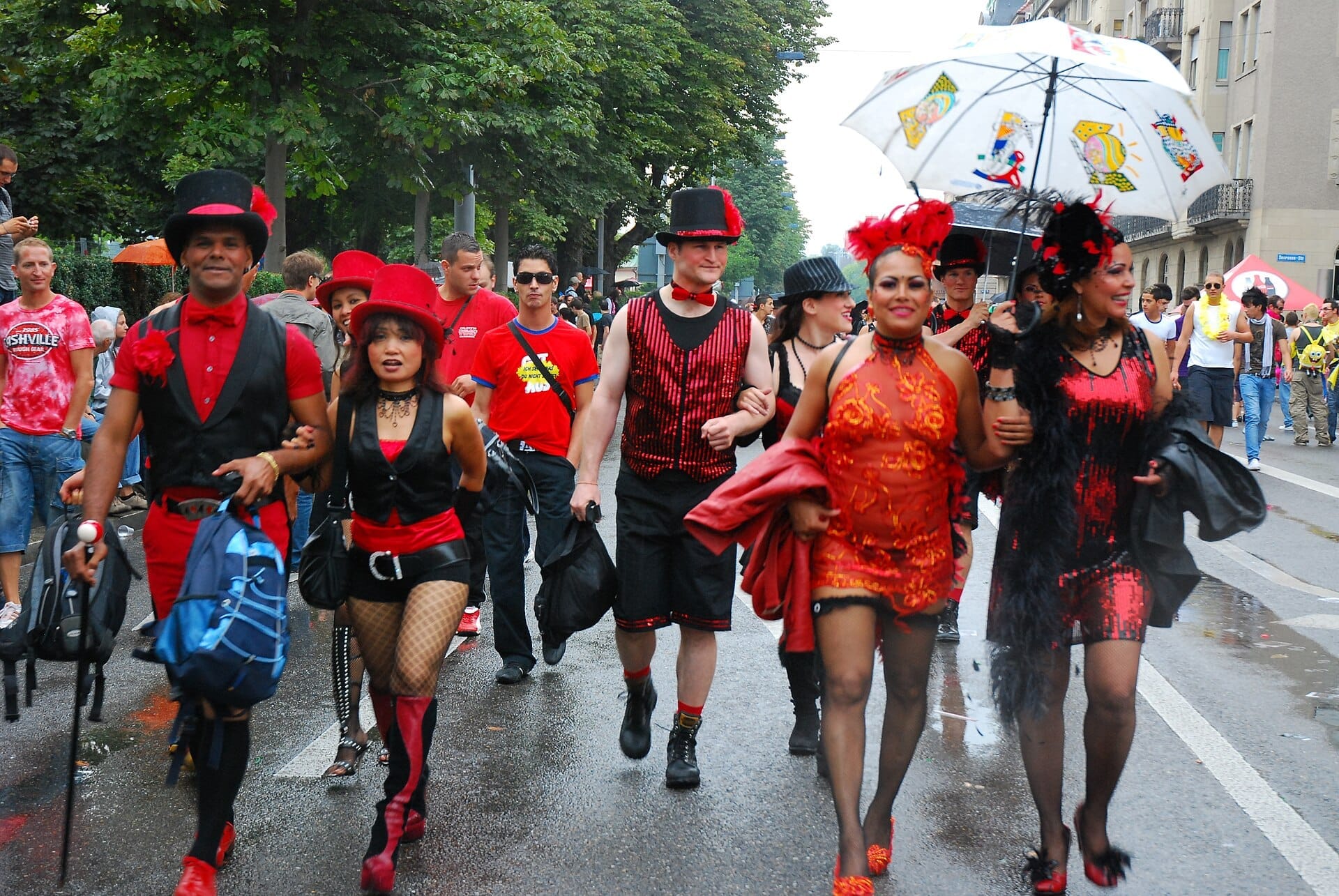 กลุ่มคนสีสันสดใสสวมชุดแดงสดใสเดินขบวนในถนนของเมืองระหว่าง Zurich Street Parade โดยมีต้นไม้และอาคารเรียงรายตลอดเส้นทาง