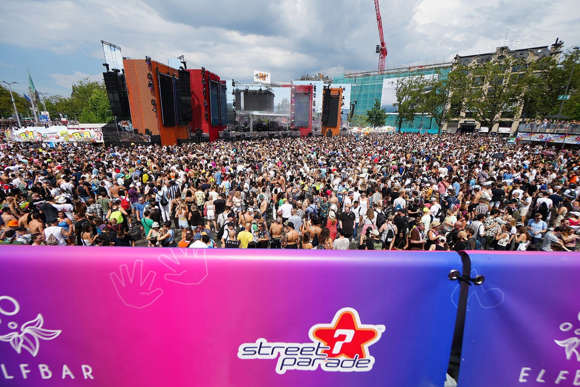 ฝูงชนจำนวนมากรวมตัวกันหน้าเวทีอันสดใสใน Zurich Street Parade พร้อมป้ายสีสันสดใส อาคารในเมือง และท้องฟ้าที่มีเมฆปกคลุมเป็นฉากหลัง