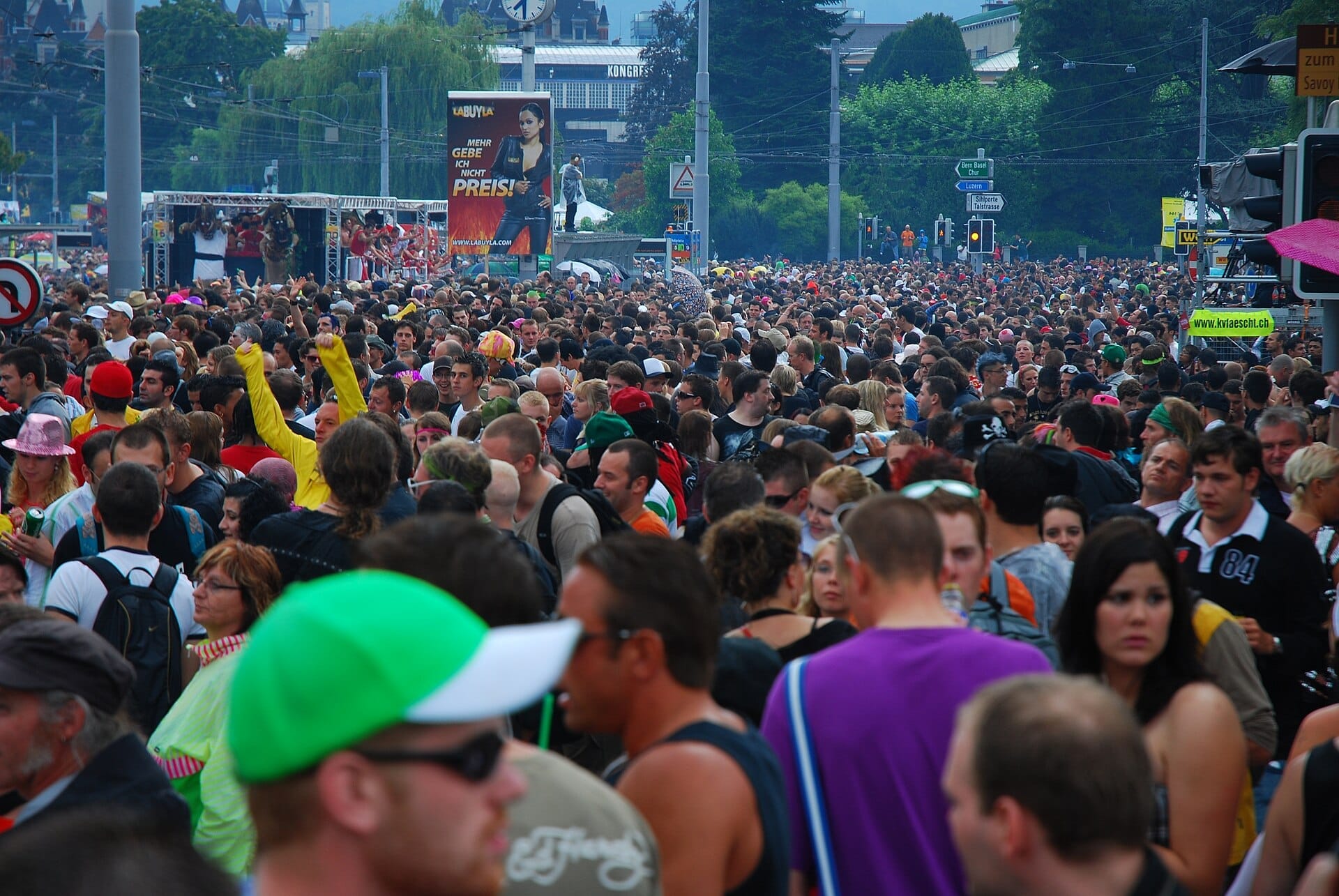 ฝูงชนจำนวนมากเต็มท้องถนนในงาน Zurich Street Parade พร้อมด้วยชุดแต่งกายสีสันสดใส ป้ายถนน และต้นไม้ที่มองเห็นได้ในพื้นหลัง