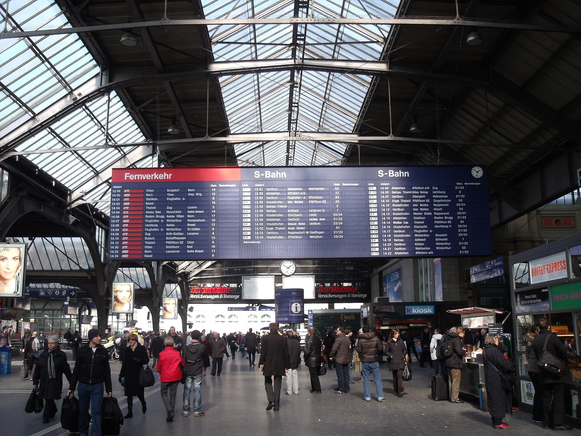 ห้องโถงที่คึกคักของ Zürich Hauptbahnhof พร้อมป้ายตารางเวลาขนาดใหญ่และนักเดินทางใต้หลังคากระจก
