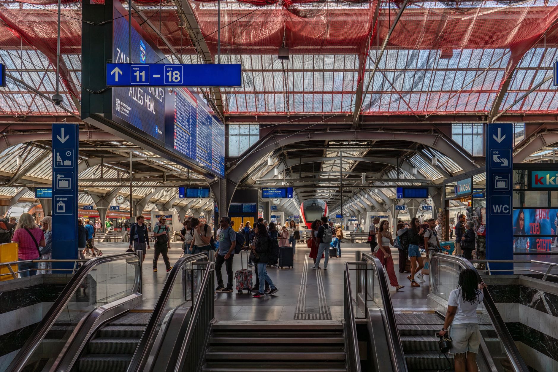 ภายในสถานีรถไฟ Zurich HB พร้อมผู้คน บันไดเลื่อน ป้ายสีน้ำเงินบอกชานชาลา และหลังคากระจก