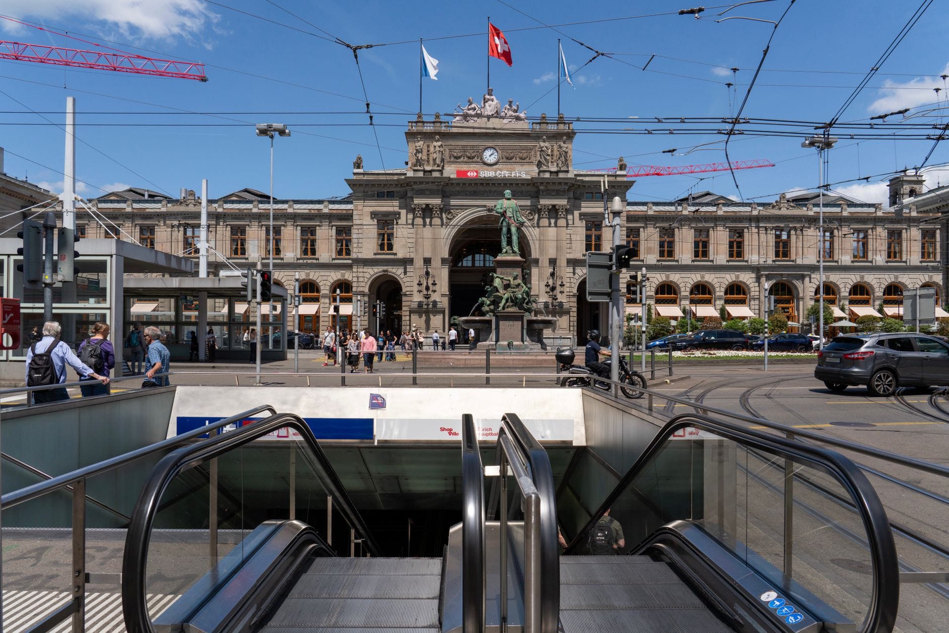 มุมมองด้านหน้าของ Zürich Hauptbahnhof พร้อมสายรถราง รูปปั้น และผู้คนในลานกว้างในวันที่แดดจ้า