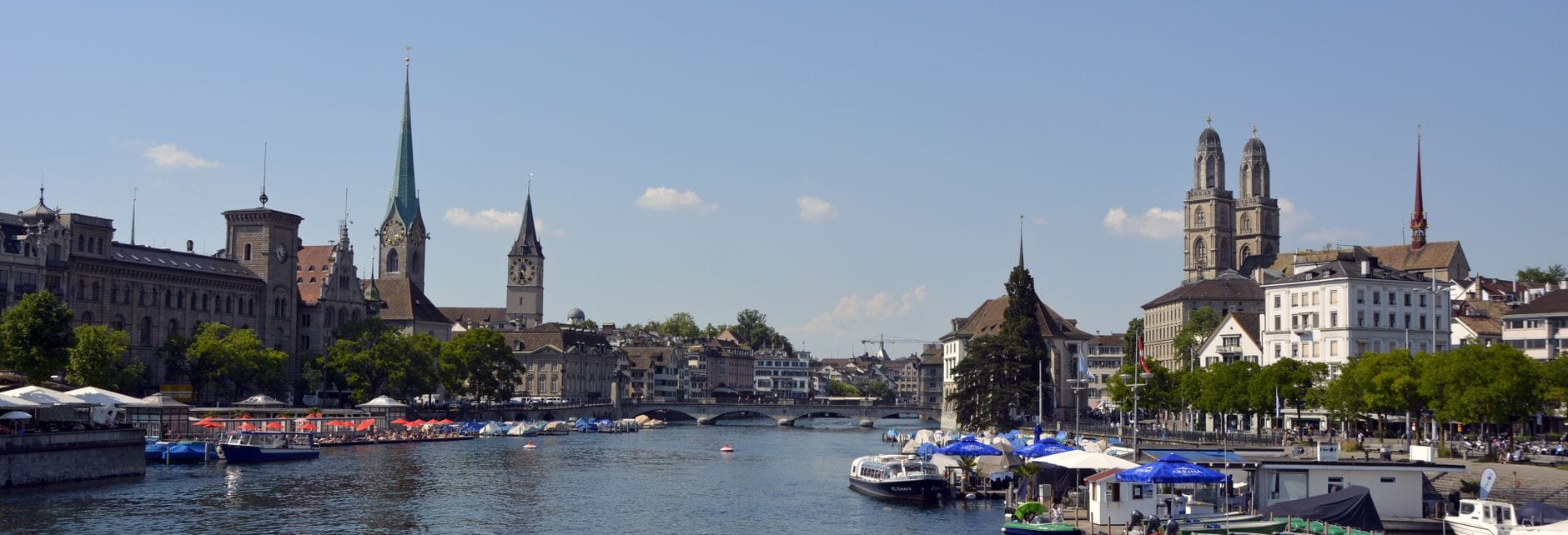 วิวพาโนรามากว้างของเมืองเก่าซูริคริมแม่น้ำ Limmat เห็นอาคารประวัติศาสตร์ ยอดหอคอย Grossmünster และเรือที่จอดตามแนวริมน้ำ ท่ามกลางท้องฟ้าโปร่งใส