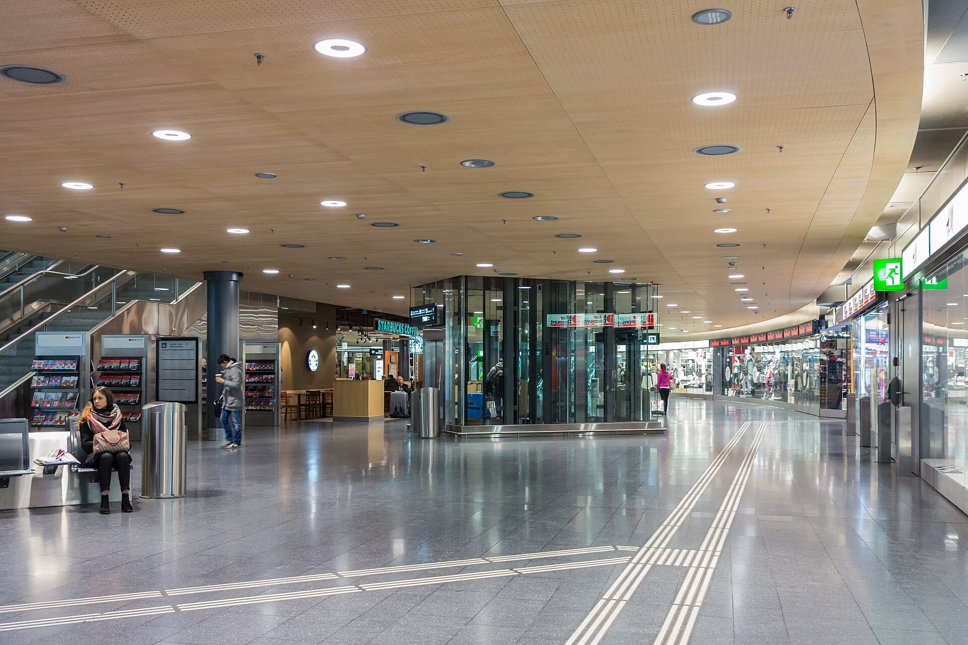 มุมมองกว้างของภายในอาคารผู้โดยสารสนามบิน Zurich Airport พร้อมร้านค้า ลิฟต์กระจก แสงไฟสมัยใหม่ และนักเดินทางที่กำลังเดินหรือนั่ง