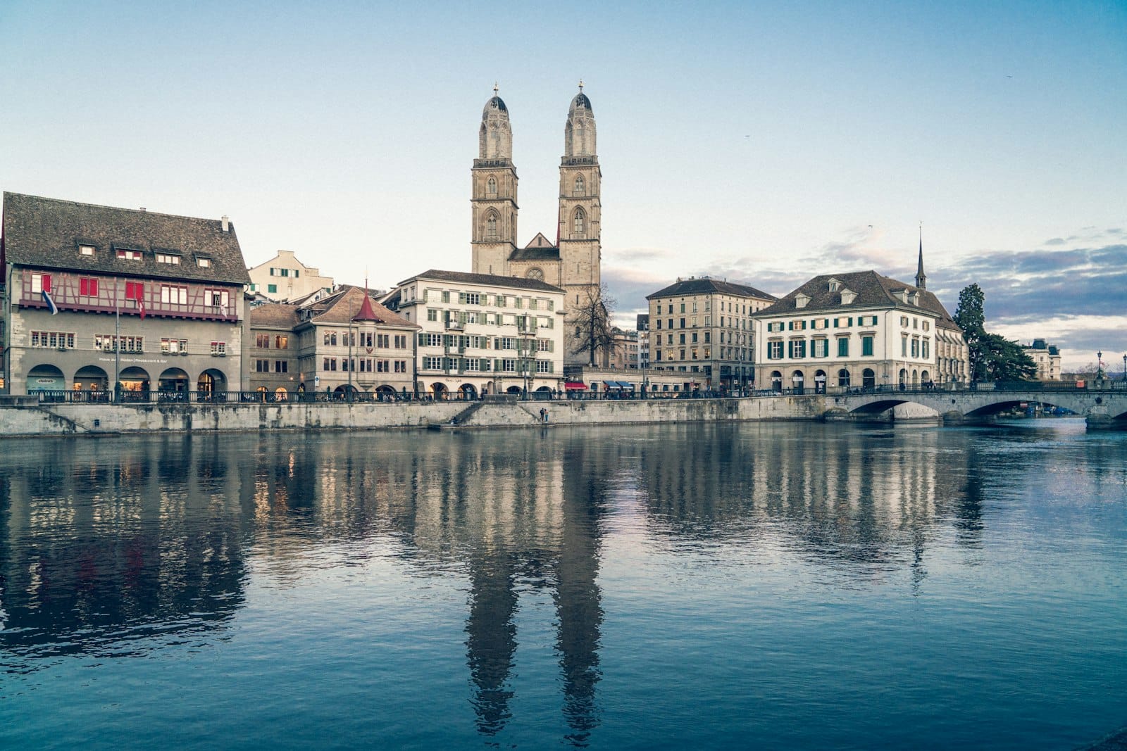 Zurich cityscape featuring the Grossmünster church with its twin towers and historic buildings reflected in the Limmat River under a clear blue sky.