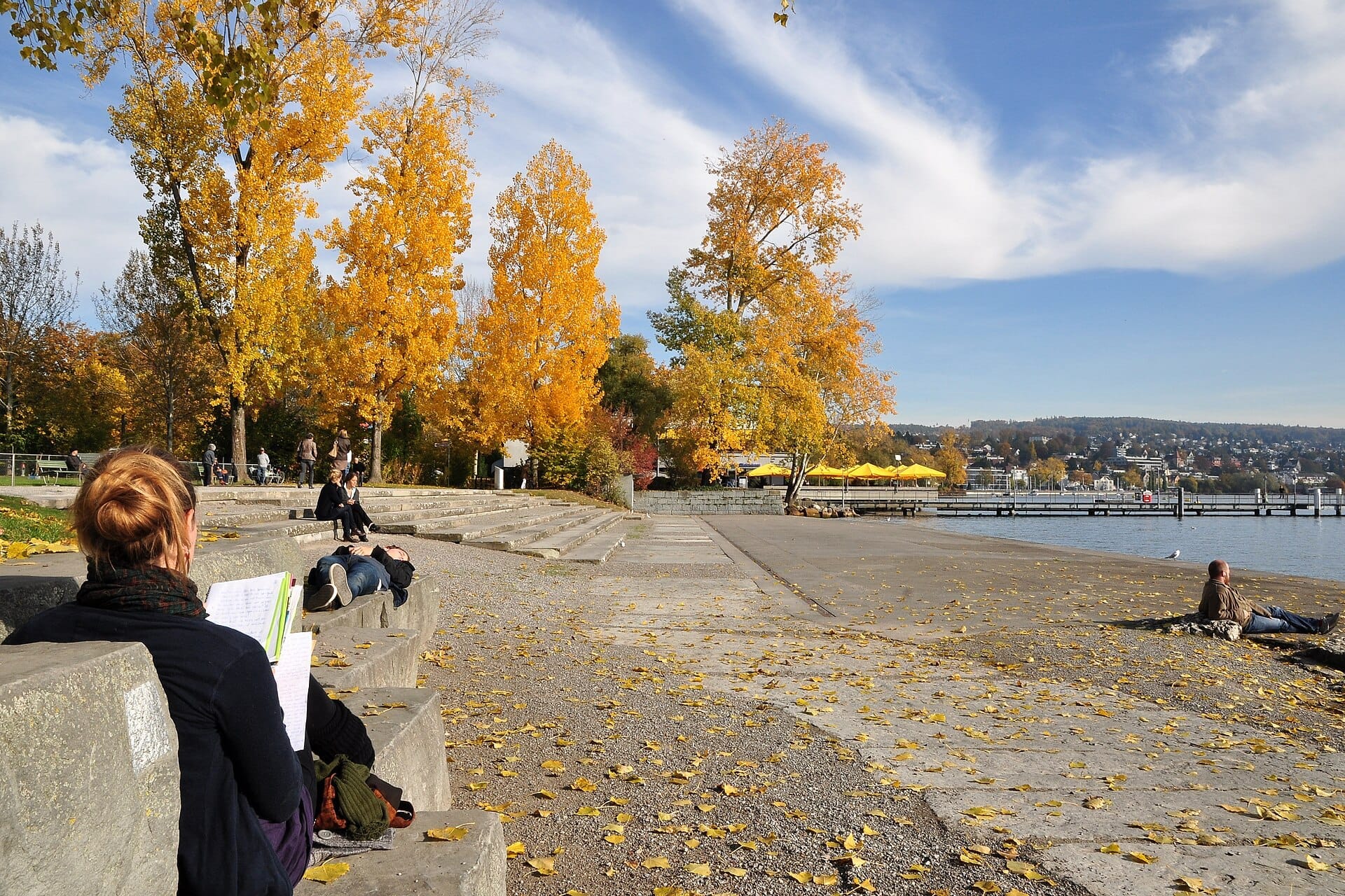 ผู้คนพักผ่อนบนบันไดหินและตามแนวชายฝั่งทะเลสาบในสวน Zürichhorn Park โดยมีต้นไม้ใบเหลืองสดใสในฤดูใบไม้ร่วงและทะเลสาบซือริชเป็นฉากหลัง